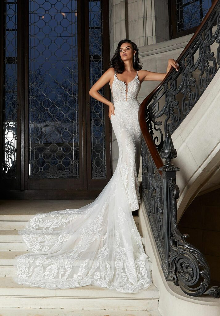 An elegant bride standing on a winding staircase in front of a window wearing a dress with a long train