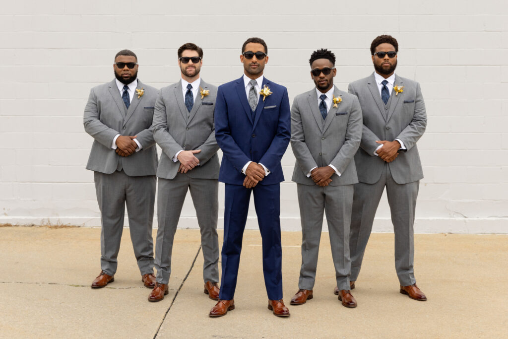 A group of men dressed in gray and navy blue tuxedos and suits, posed in front of a brick wall.