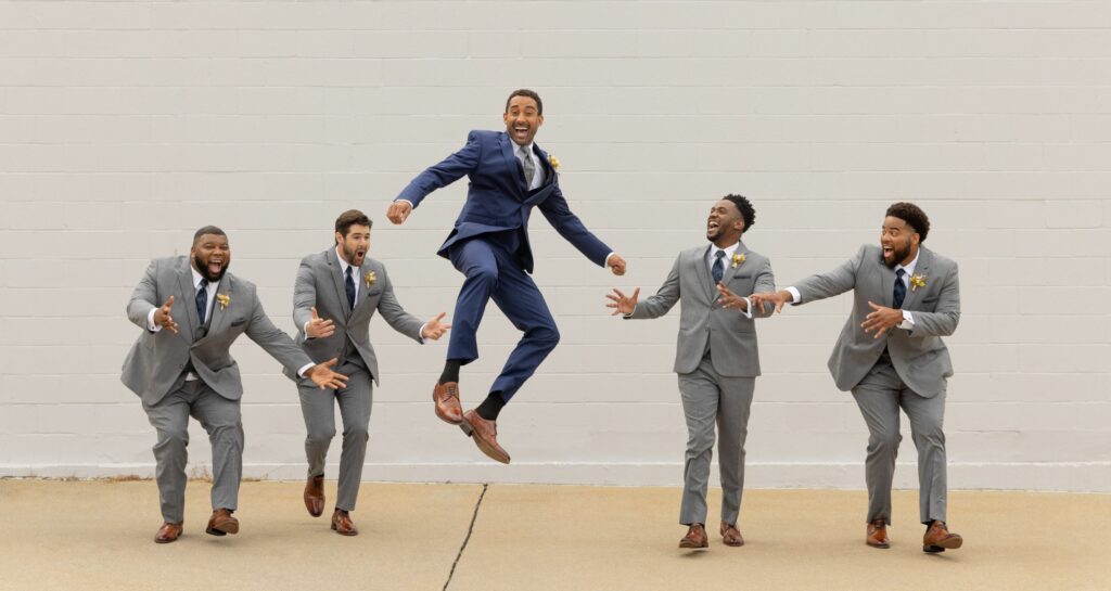 A group of four groomsmen posing around the groom who is jumping in a blue suit.