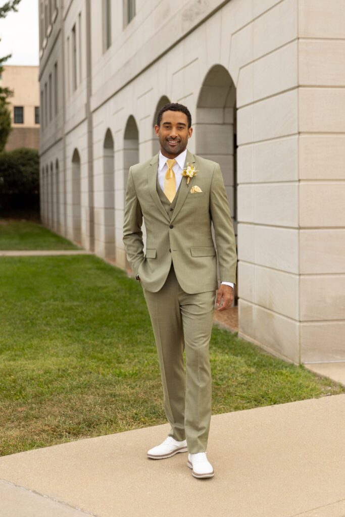 A groom dressed in a sage green suit and yellow tie standing outside the wedding venue.