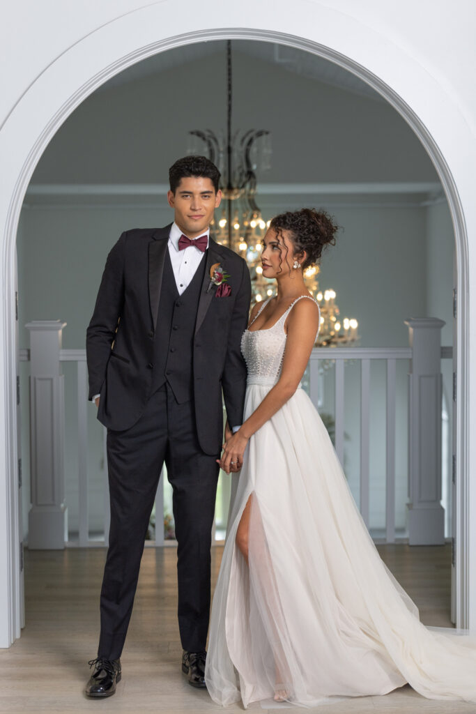 A bride and groom posing with each other in front of a balcony and chandelier, in their dress and tuxedo.