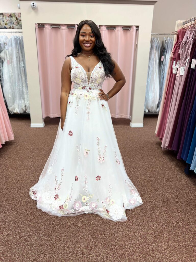 Happy bride in a floral wedding dress smiling in a bridal shop, showing off the detailed floral embroidery.