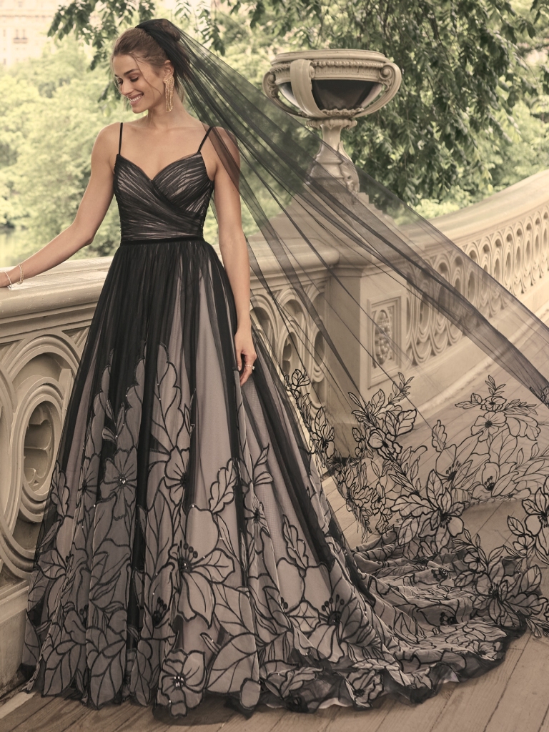 Bride in a dramatic black wedding dress with floral overlay, smiling in an urban park setting.