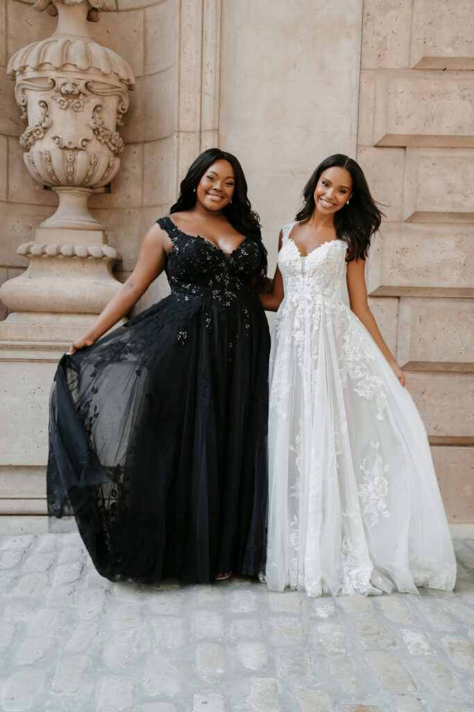 Two brides, one in a black floral gown and one in white, posing elegantly in a historic setting.