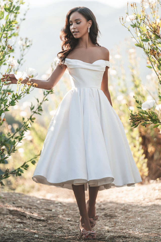 Bride in a charming white off-shoulder gown with a flowy skirt, touching flowers in a garden.