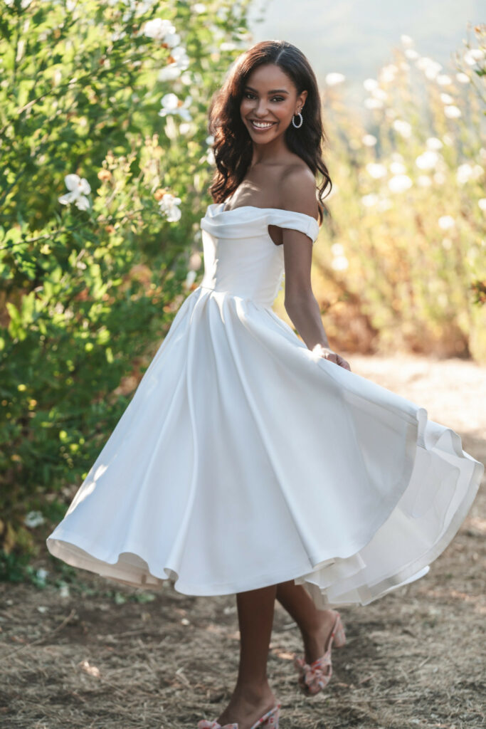 Bride in a white off-shoulder gown twirling joyfully in a sunny garden.