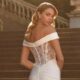 Elegant bride in a white gown with off-shoulder detail and lace back, posing on steps.