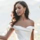 Bride in an elegant off-the-shoulder white dress surrounded by delicate flowers, embracing a serene outdoor setting.