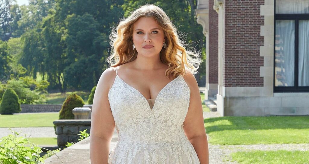 Plus-size bride in a lace wedding gown with a deep V-neckline, standing confidently in front of a grand estate.