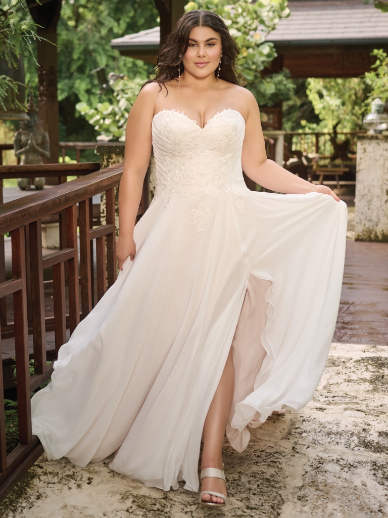 Woman in a strapless, sweetheart-neckline wedding dress with a flowy skirt, posing outdoors near a wooden railing.