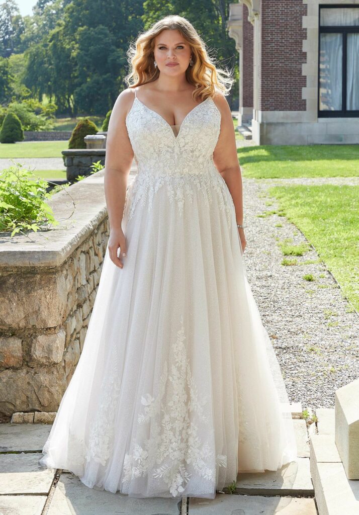 Front-facing view of a bride in a detailed lace ballgown with a V-neckline, outdoors on a sunny day near lush greenery.