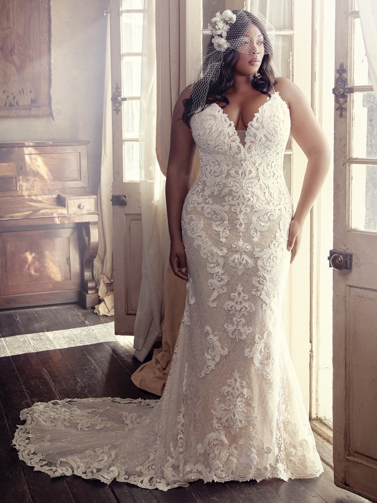 Plus-size bride in an intricately laced fit-and-flare gown, styled with a vintage veil in a softly lit rustic room.