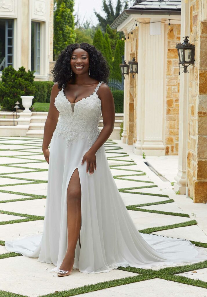 Smiling woman in a lace wedding dress with spaghetti straps and a thigh slit, posing in a luxurious outdoor setting.