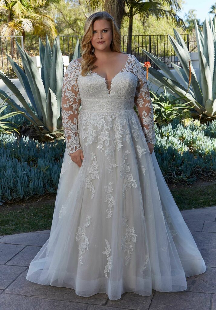 Bride in a long-sleeve A-line lace wedding gown, posing outdoors near greenery and architectural elements.