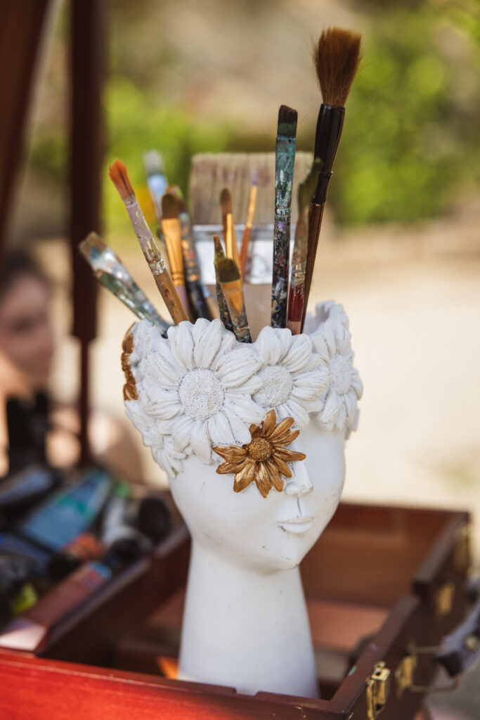 A white vase with floral embellishments holding various paintbrushes, set outdoors during a wedding event.