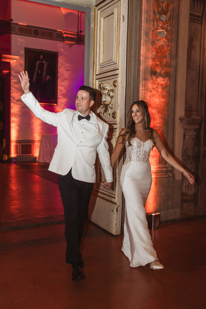 Bride and groom entering a reception hall; bride in a fitted gown with a lace bodice, groom in a white tuxedo jacket waving.