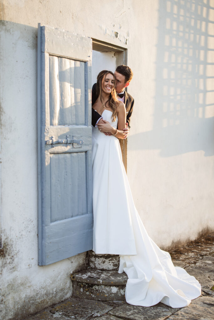 Bride leaning against a blue rustic door, her groom embracing her; both smiling warmly with golden sunlight in the background.