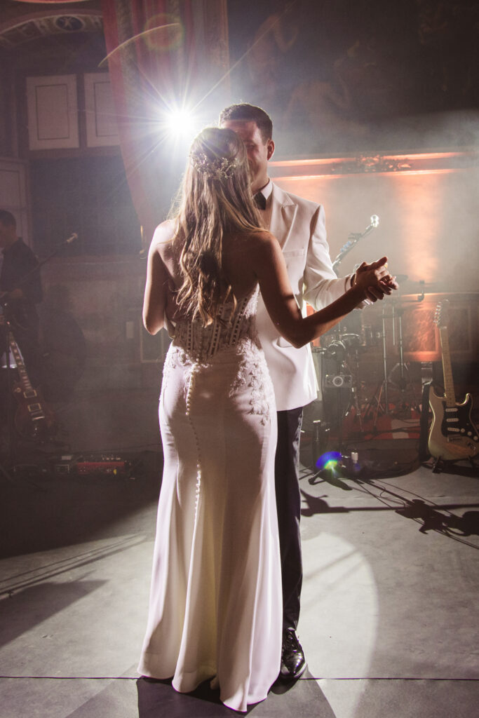 A couple shares their first dance under dramatic lighting, the bride in a fitted gown and the groom in a white suit.