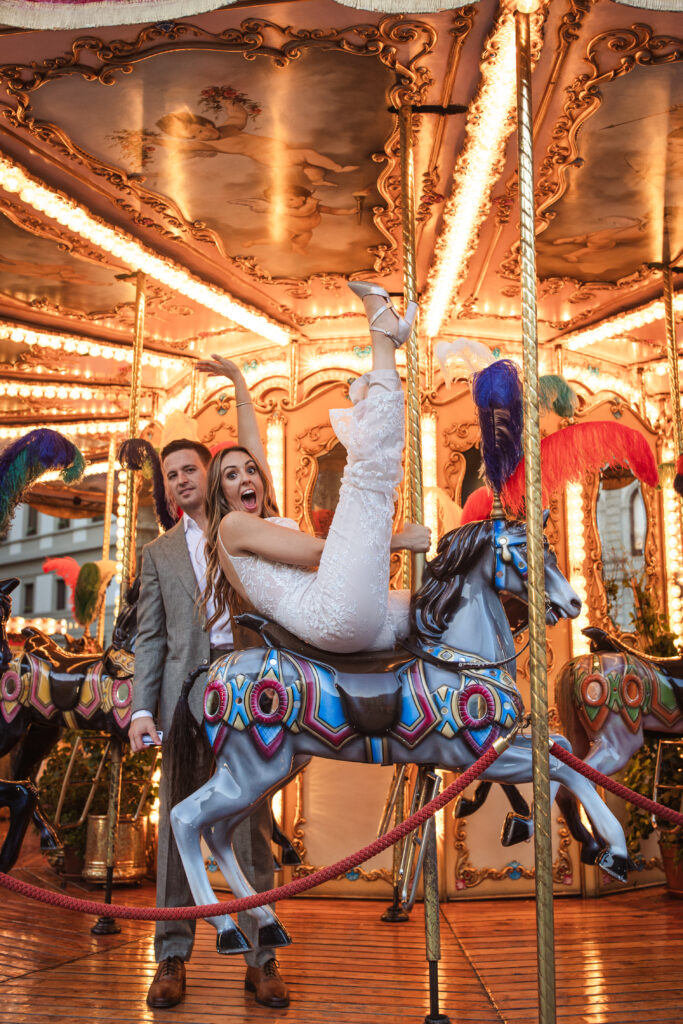 Bride playfully posing on a carousel horse, wearing a lace jumpsuit with intricate detailing, alongside her smiling groom.