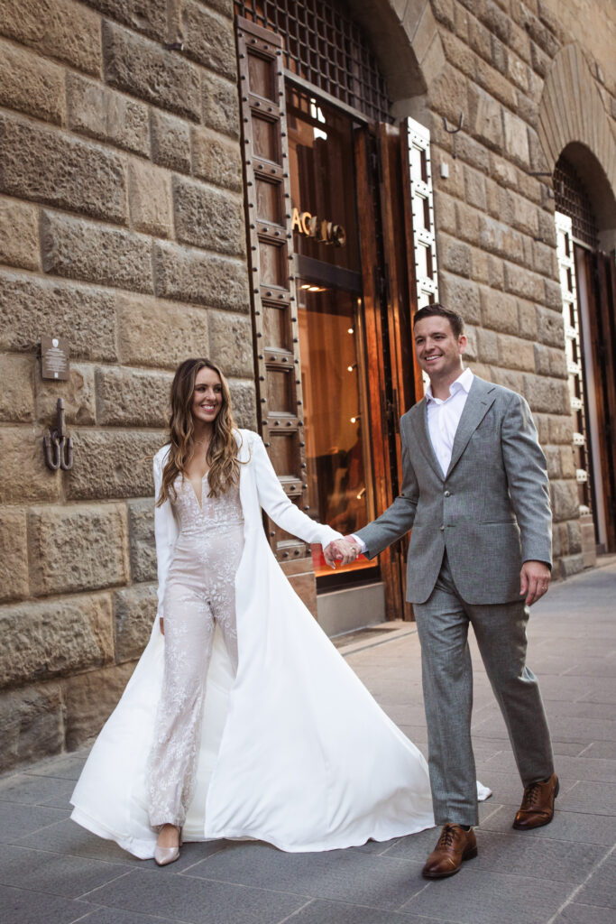 Couple walking hand-in-hand in a cityscape; bride in a lace jumpsuit with a dramatic white overcoat, groom in a gray suit.