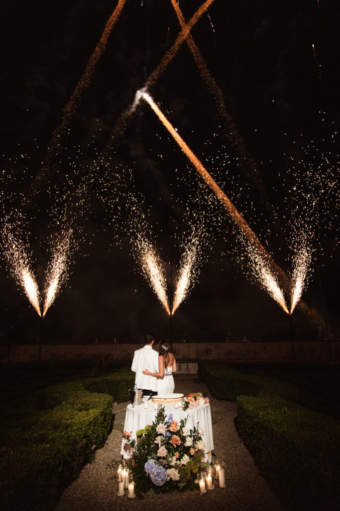 A romantic wedding setting with a table adorned with floral arrangements and lit candles, framed by fireworks.