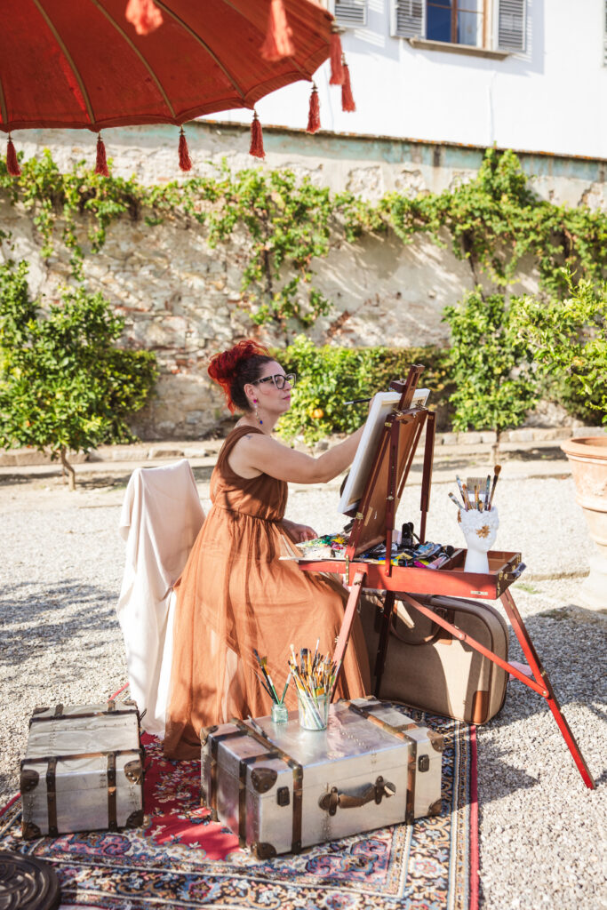 A painter in a burnt orange dress captures a wedding scene, sitting beside vintage trunks and art supplies.