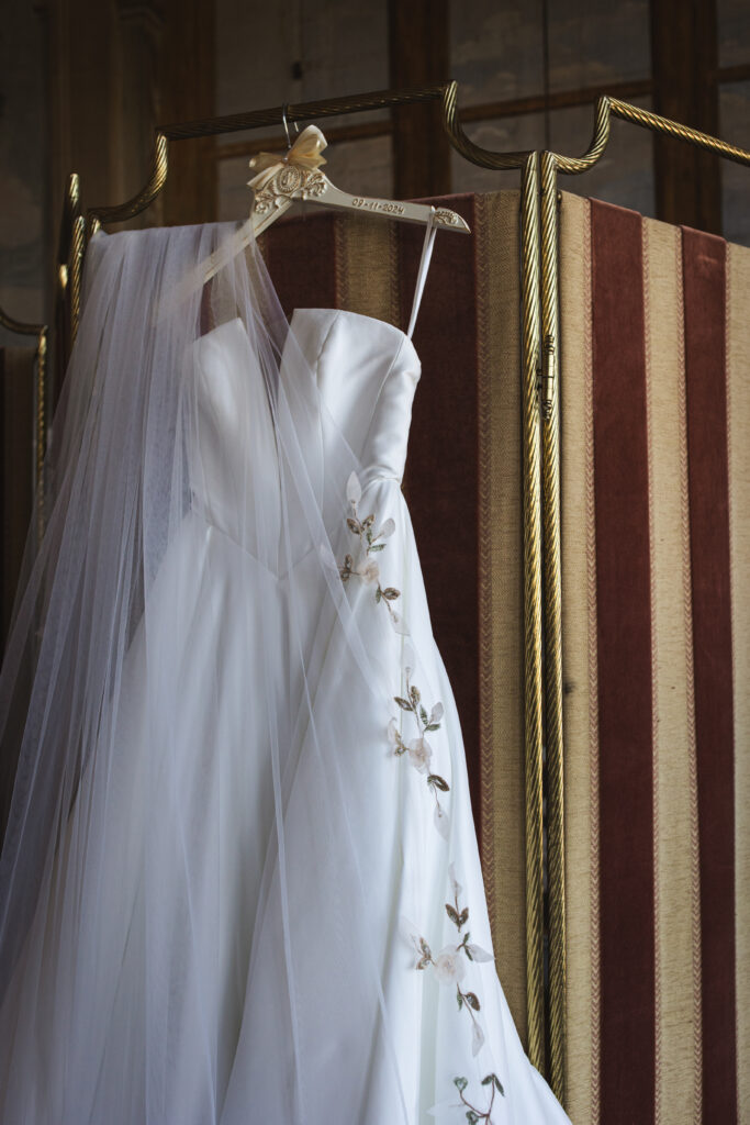 Elegant wedding dress with floral appliqués displayed on a gold hanger next to a striped background and a flowing veil.