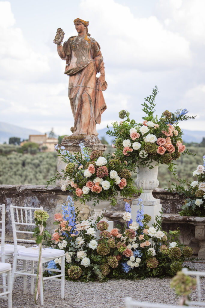 A Tuscan-inspired wedding setup featuring a rustic statue surrounded by lush floral arrangements in peach and white.