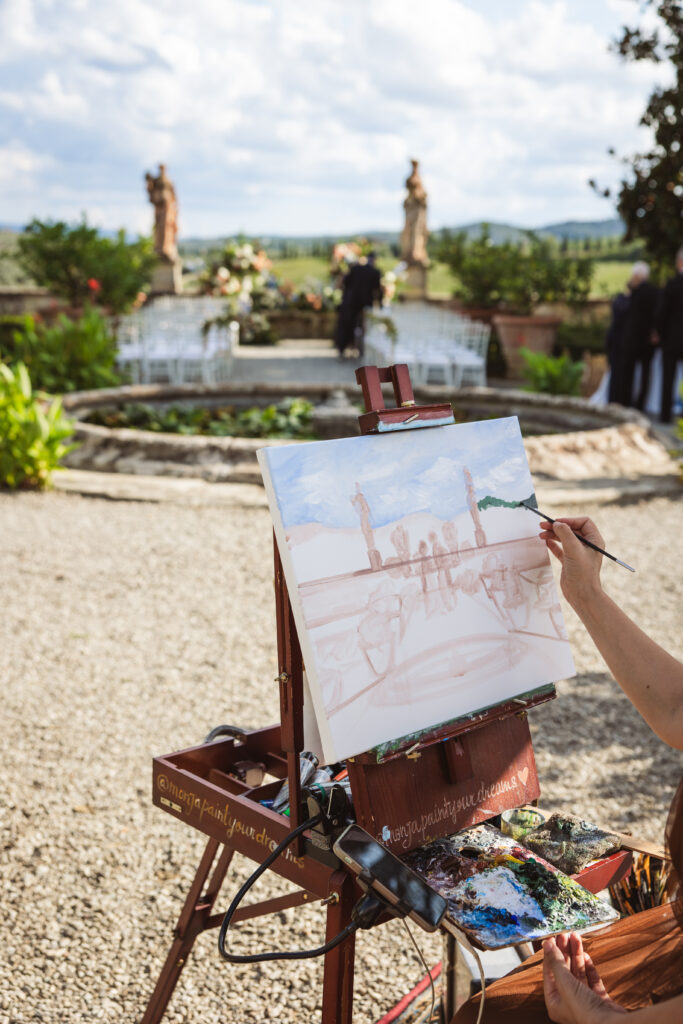 An easel displaying a live painting of a wedding ceremony, with a scenic backdrop of a vineyard and statues.