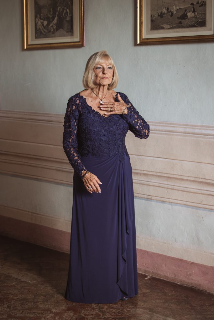 Woman poses in a navy lace gown with a ruched skirt, standing in a vintage room featuring framed artwork on the walls.