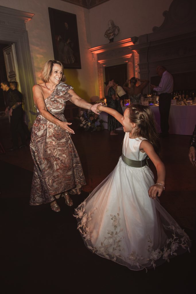 A mother-of-the-bride dances joyfully with a young flower girl in a white dress with floral embellishments at a wedding reception.