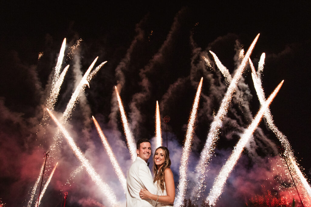 A newlywed couple smiling with vibrant fireworks illuminating the night sky behind them.