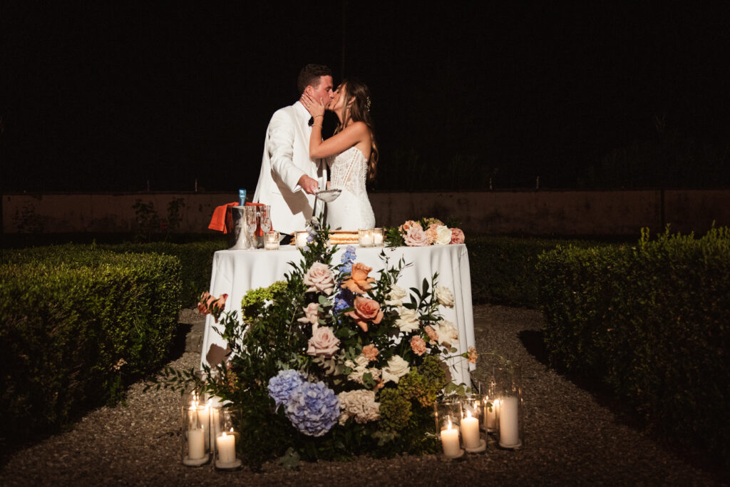 Romantic nighttime moment of the bride and groom sharing a kiss by a candlelit wedding cake adorned with flowers.