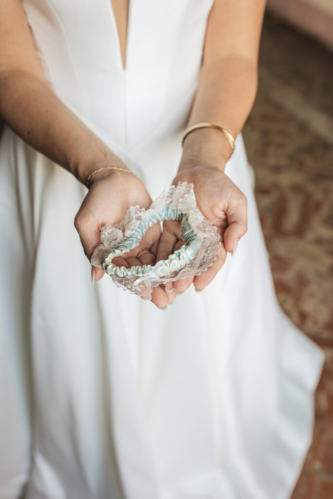 Bride holding a delicate lace garter in her hands, showcasing intricate details of her white gown with a plunging neckline.