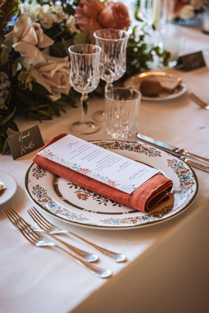 Close-up of ornate wedding dinnerware and a menu accented by burnt orange napkins and intricate floral designs.