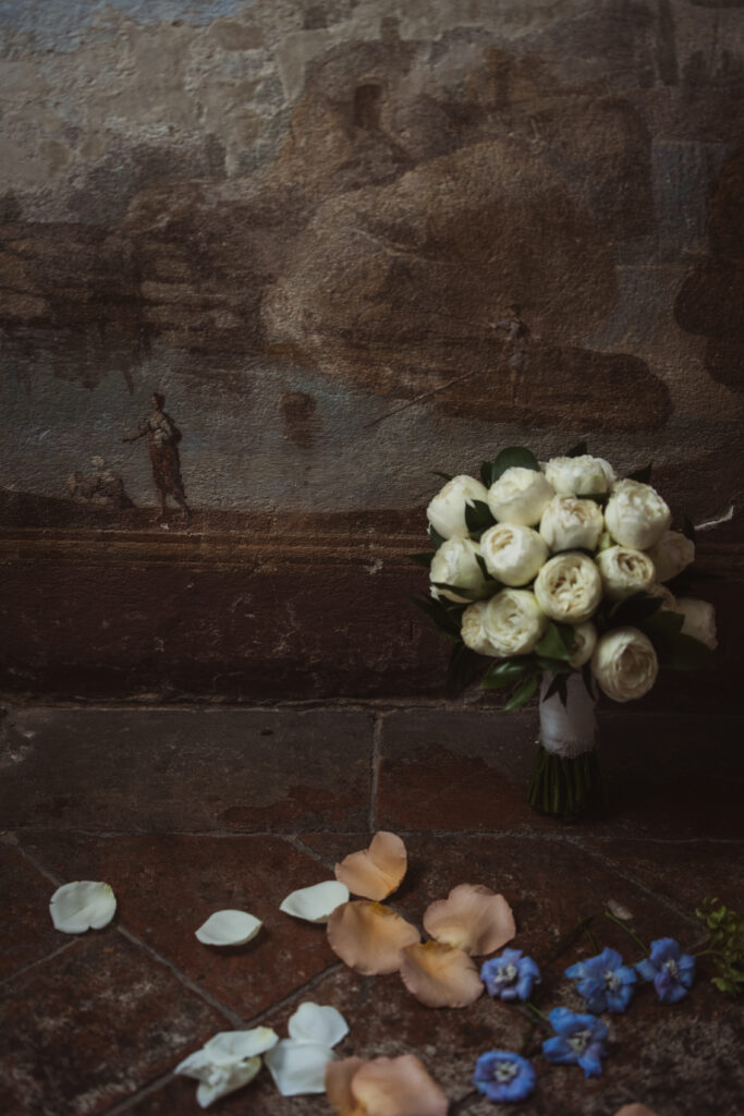 White rose bridal bouquet placed against an antique Italian fresco wall with scattered peach and blue flower petals.