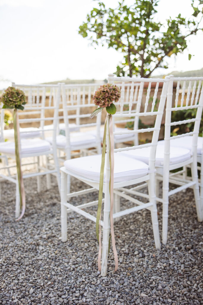 Rows of white chairs adorned with hydrangea bouquets and pastel ribbons, set on gravel at a scenic outdoor wedding.