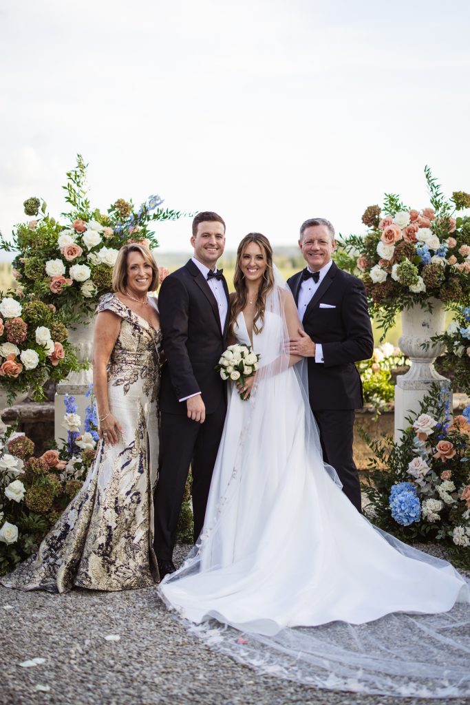 A family portrait of the bride, groom, and parents in formal attire, standing by floral arrangements under a clear sky.