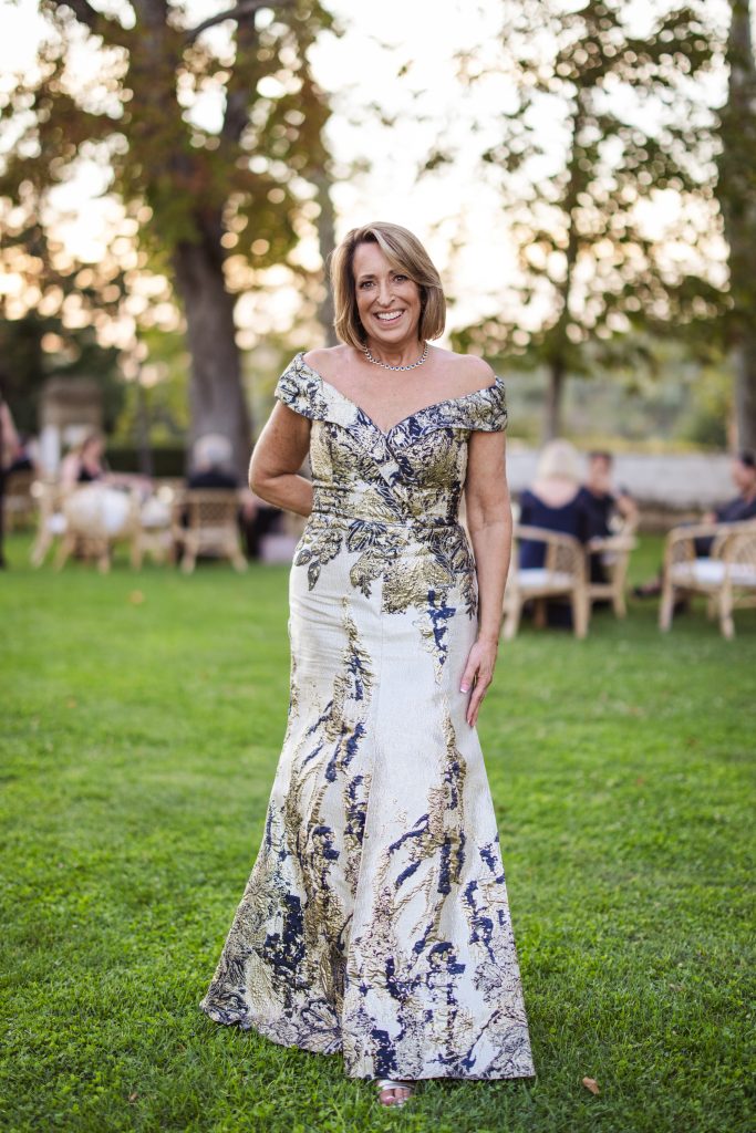 A mother-of-the-bride in a gold and blue gown smiles gracefully while standing on a green lawn with trees in the background.