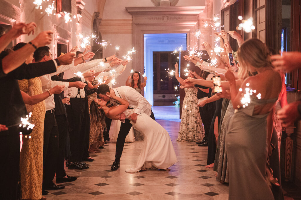 Bride and groom kissing surrounded by guests holding sparklers in a grand Italian villa hallway.