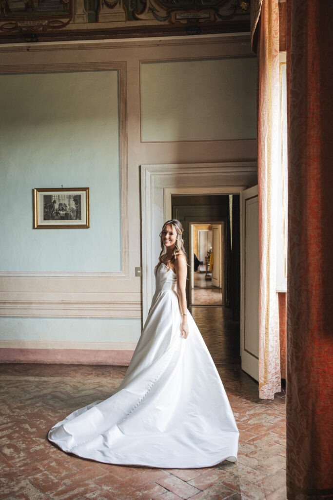 Bride standing in a vintage-inspired room with high ceilings and soft lighting, wearing a satin ballgown with button details.