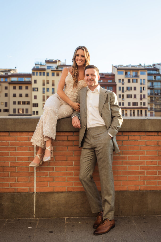 Bride and groom posed outdoors; bride in an intricately detailed lace jumpsuit, groom in a tailored gray suit, buildings in the backdrop.