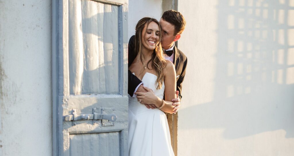 Bride in a strapless ball gown smiling while embracing the groom, framed by a rustic blue door in a soft-lit setting.