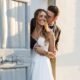 Bride in a strapless ball gown smiling while embracing the groom, framed by a rustic blue door in a soft-lit setting.