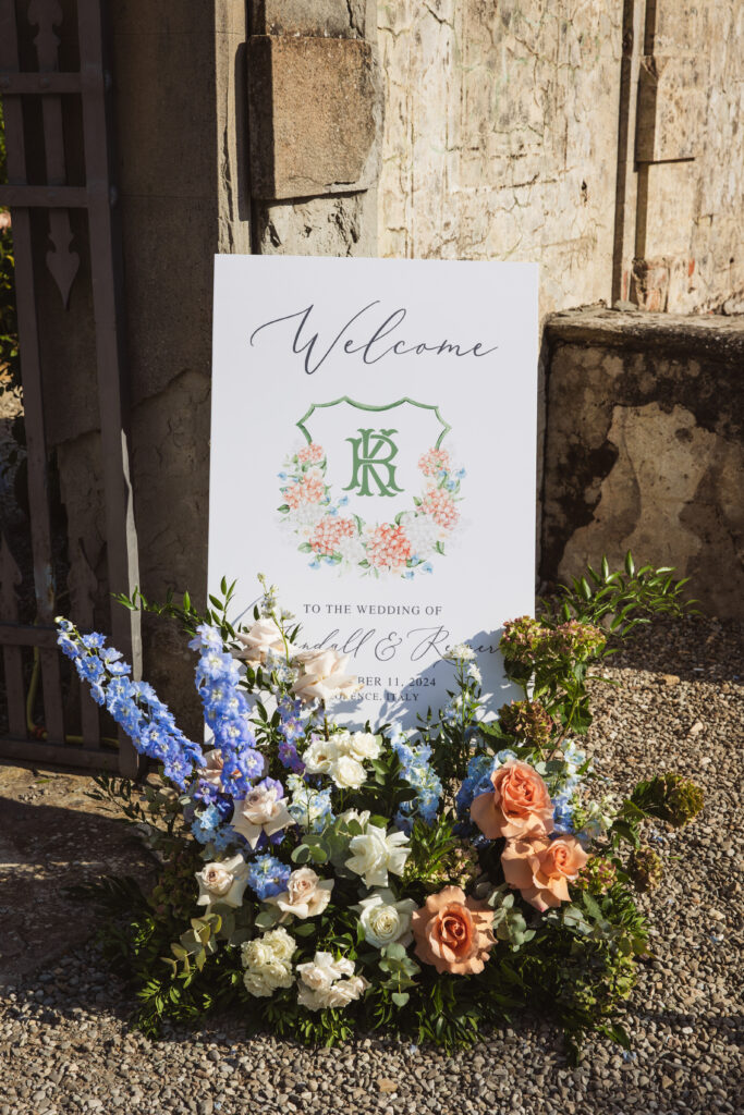 A beautifully designed wedding welcome sign surrounded by lush floral arrangements at an Italian villa.