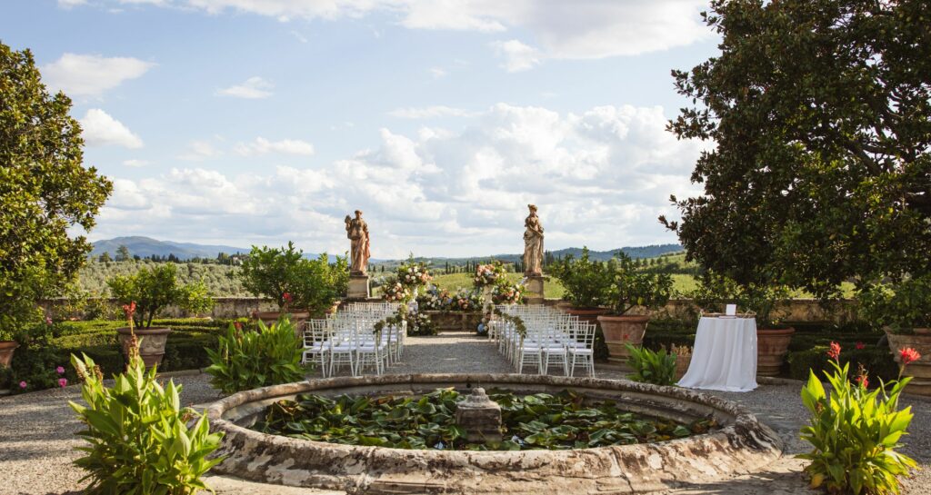 Outdoor wedding ceremony setup in an Italian villa garden, featuring a lily pond, floral arrangements, and scenic countryside views.