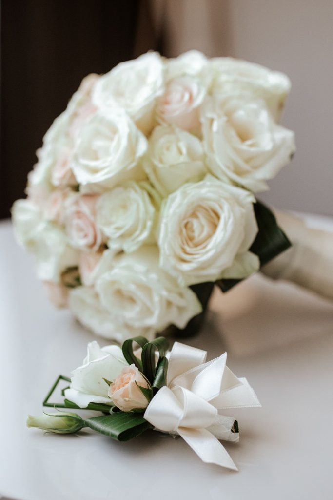 A timeless wedding bouquet of white roses, accompanied by a matching boutonniere with soft blush roses and a white ribbon, elegantly displayed on a table.