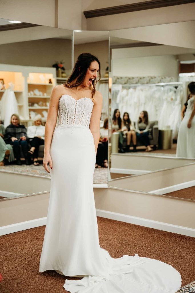 A bride-to-be at Aurora Bridal excitedly modeling a strapless wedding gown with a lace corset bodice and a flowing white train in a bridal boutique.