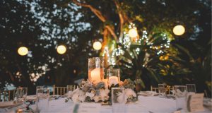 A romantic wedding reception table set outdoors at nighttime, featuring candlelit centerpieces, greenery, and glowing string lights in the background.