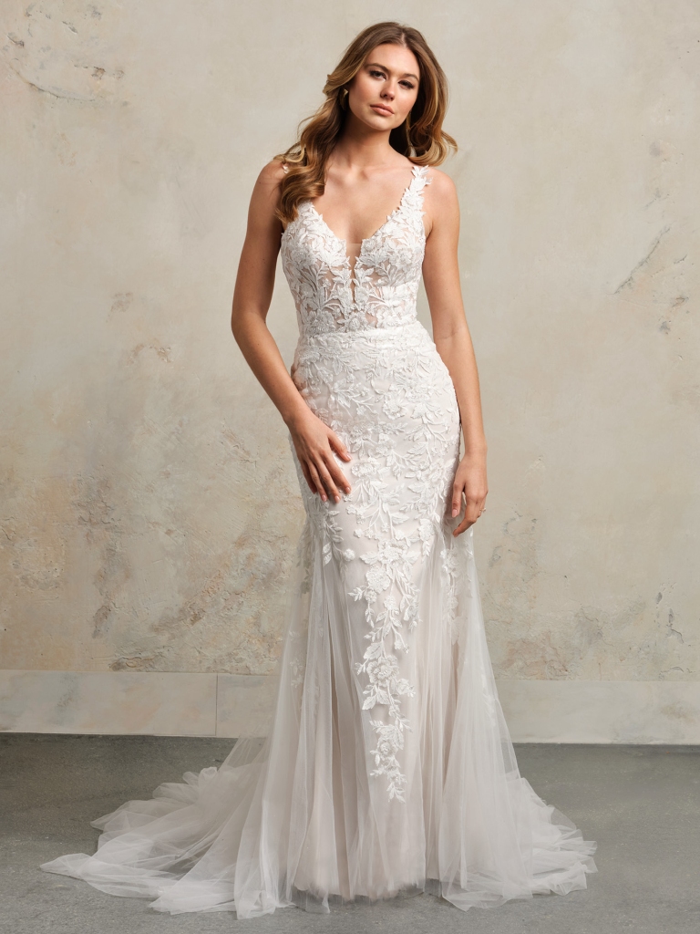 A bride wears a romantic fitted gown with leaf-patterned lace and illusion detail, creating a soft, feminine look for a beach or garden wedding.